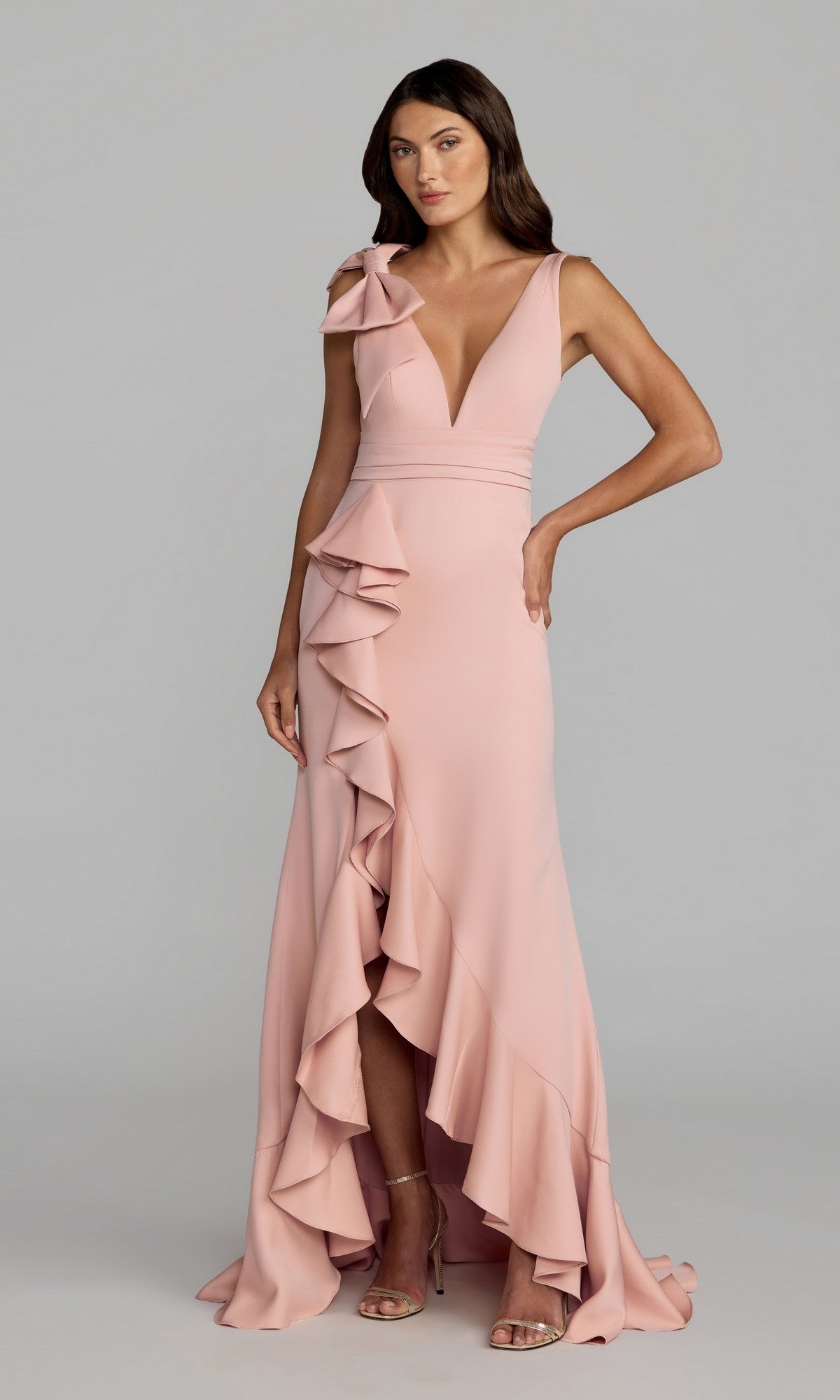A woman wearing the Mac Duggal 49965 long formal dress in blush, featuring a sleeveless design with a deep V-neck, ruffle details on the skirt, and a bow accent on one shoulder, stands against a plain light background.